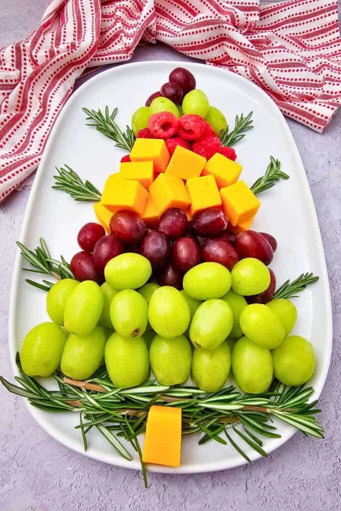 Christmas tree fruit tray arranged on a white platter with layers of green and red grapes, cheddar cheese cubes, fresh raspberries, and rosemary sprigs forming festive tree branches.