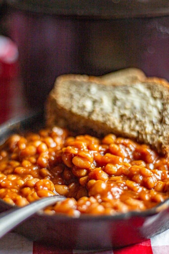 Close-up of homemade baked beans in a cast iron skillet with a rich, thick sauce. Two buttered slices of rustic bread rest at the edge of the skillet, ready for dipping.