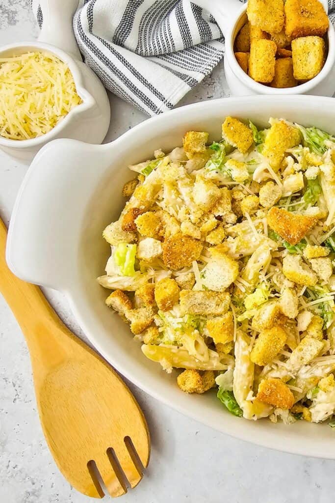 Homemade Caesar pasta salad in a white serving dish topped with crispy croutons, shaved Parmesan, and chopped romaine lettuce, with a wooden serving spoon and small bowls of cheese and croutons nearby ready for serving.