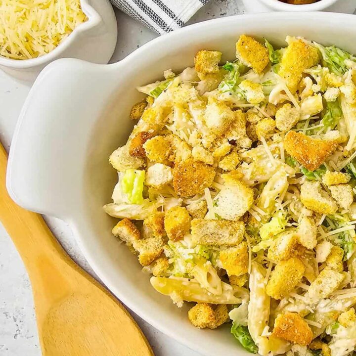 Homemade Caesar pasta salad in a white serving dish topped with crispy croutons, shaved Parmesan, and chopped romaine lettuce, with a wooden serving spoon and small bowls of cheese and croutons nearby ready for serving.