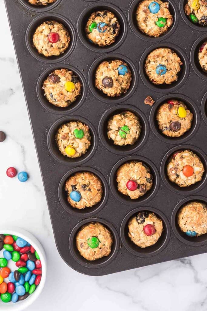 Mini baked granola cups in a muffin tin, made with oats and studded with colorful chocolate candies. A bowl of extra candy-coated chocolates sits nearby on the marble countertop.