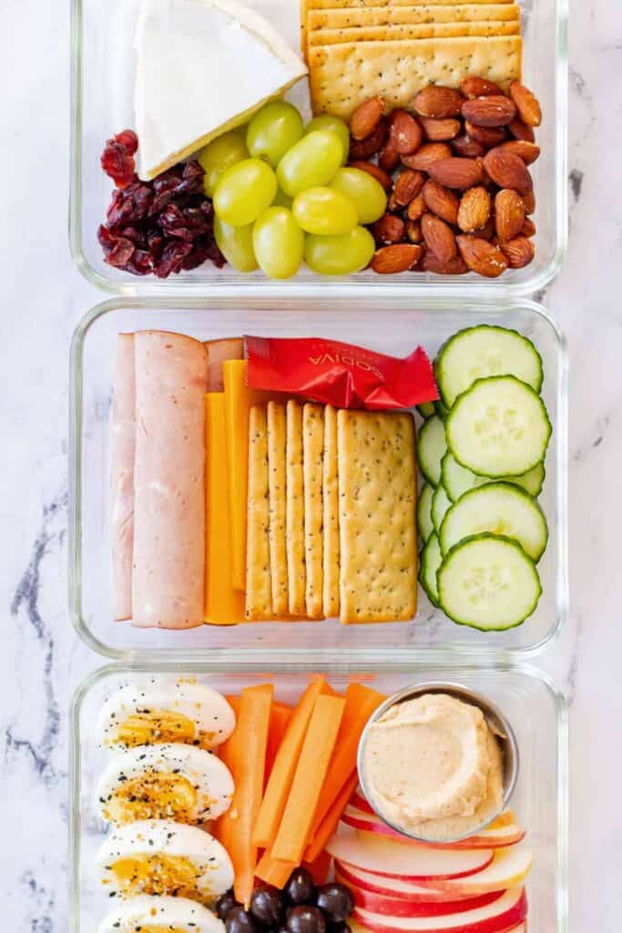 hree glass containers arranged with homemade lunchables including crackers sliced cheese and deli meat cucumber slices almonds grapes dried cranberries and a wedge of soft cheese. The bottom container shows hard boiled eggs carrot sticks apple slices hummus and olives creating a balanced set of on the go snacks.