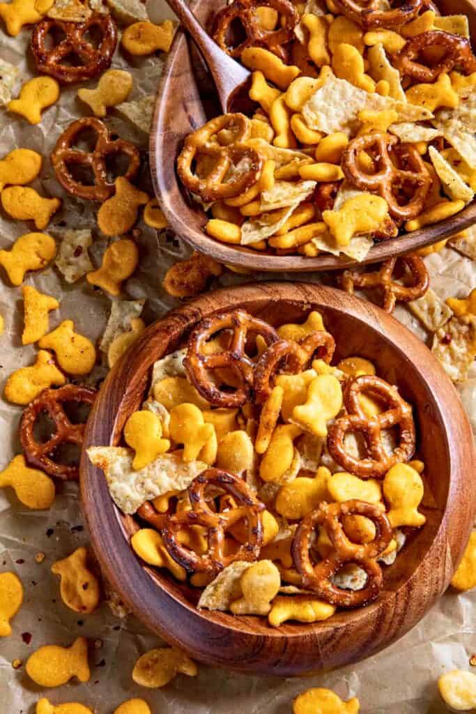 Two wooden bowls filled with homemade spicy snack mix made with pretzels cheese crackers fish shaped crackers and seasoned chip pieces scattered on parchment paper. Extra pieces are spread around the bowls showing the crunchy texture and bold seasoning of the mix.
