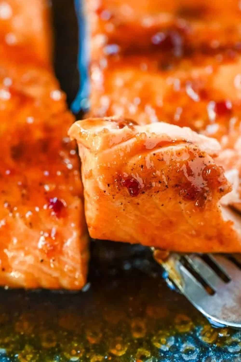 Close up of a fork lifting a flaky piece of honey garlic salmon coated in a glossy glaze with visible garlic and red pepper flakes. The caramelized salmon is shown fresh from the pan, highlighting its tender texture and sticky sweet savory sauce.