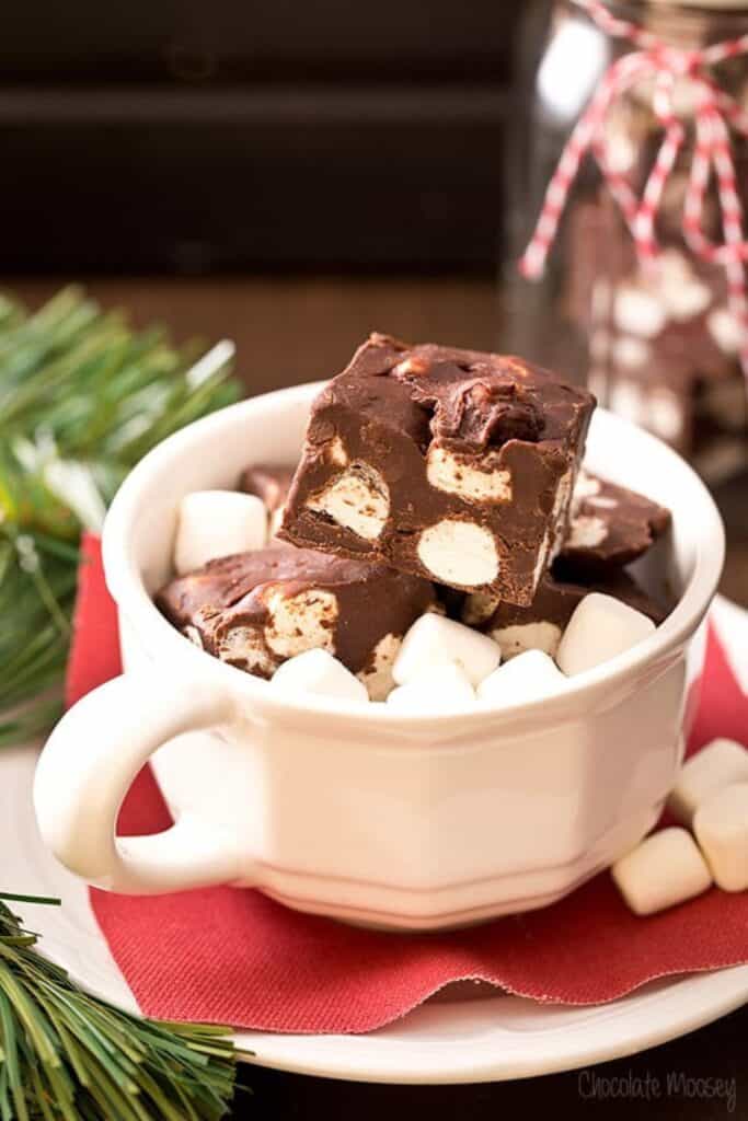 Squares of hot chocolate fudge filled with mini marshmallows and chocolate chunks, served in a white mug with extra marshmallows. Set on a red napkin with holiday greenery, creating a cozy and festive holiday presentation.