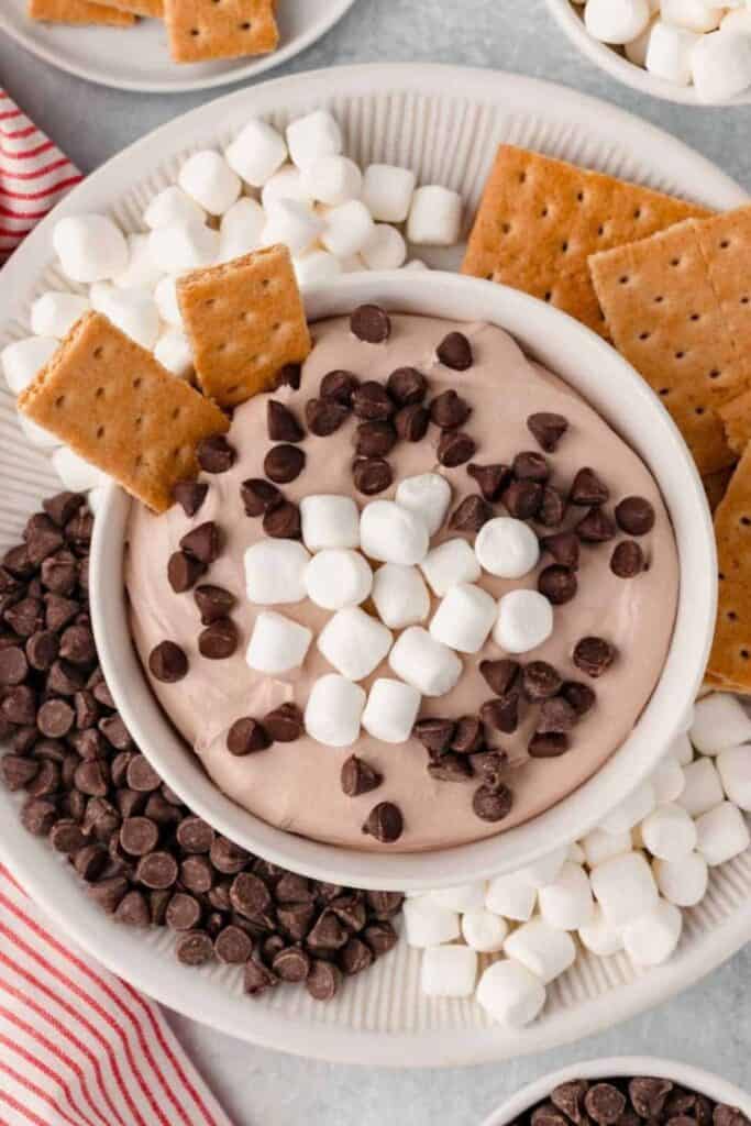 A bowl of creamy hot cocoa dip topped with mini marshmallows and chocolate chips, surrounded by graham crackers and more marshmallows for dipping, served on a festive tray.