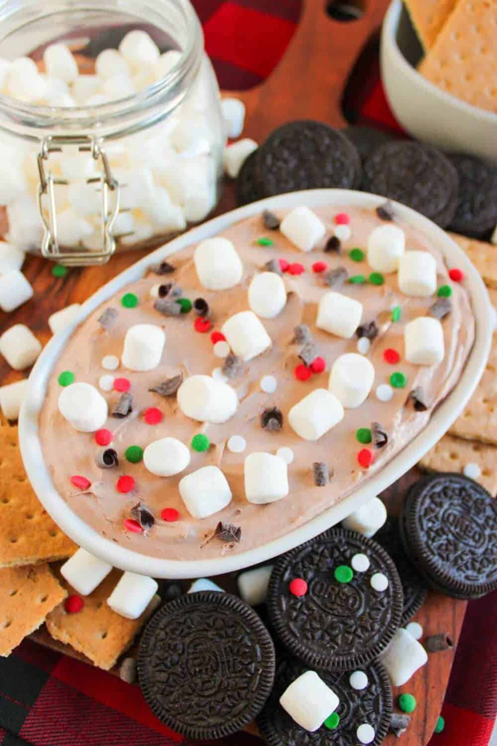 Creamy hot cocoa dip topped with mini marshmallows, chocolate curls, and red, green, and white sprinkles. The festive dessert dip is surrounded by chocolate sandwich cookies, graham crackers, and extra marshmallows for dipping.