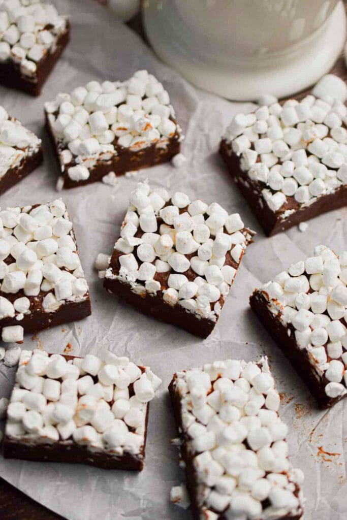 Squares of rich hot cocoa fudge topped with a generous layer of mini marshmallows, arranged on parchment paper. The soft, fluffy topping adds a cozy, wintery feel perfect for holiday treats.