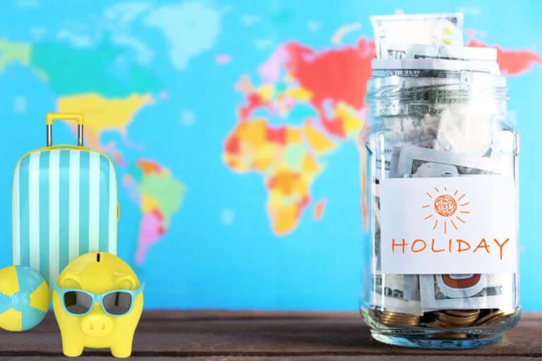 A glass jar labeled “Holiday” filled with U.S. dollar bills and coins sits in front of a colorful world map, symbolizing saving money for travel. Beside it are a striped blue suitcase, a yellow piggy bank wearing sunglasses, and a beach ball, reinforcing the vacation theme.
