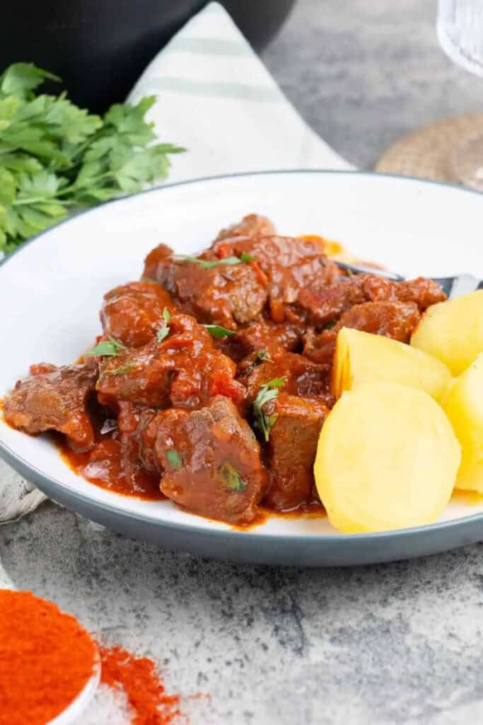 Round plate filled with Hungarian beef stew with pieces of beef in sauce, beside boiled potatoe chunks. A small dish of Hungarian paprika sits in front.