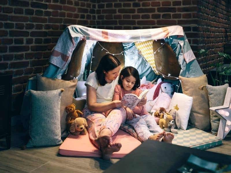 Mom and daughter camping indoors reading a book. 