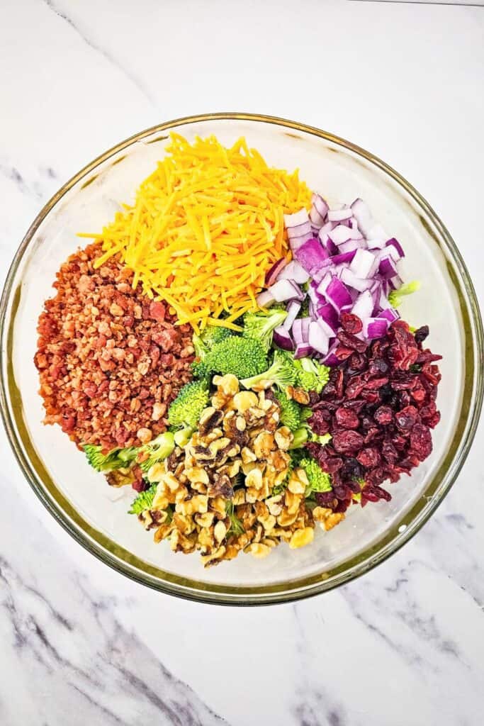 Overhead view of a large glass bowl filled with individual piles of salad ingredients: shredded cheddar cheese, diced red onion, dried cranberries, chopped walnuts, fresh broccoli florets, and crispy bacon bits, ready to be mixed into a broccoli pasta salad.