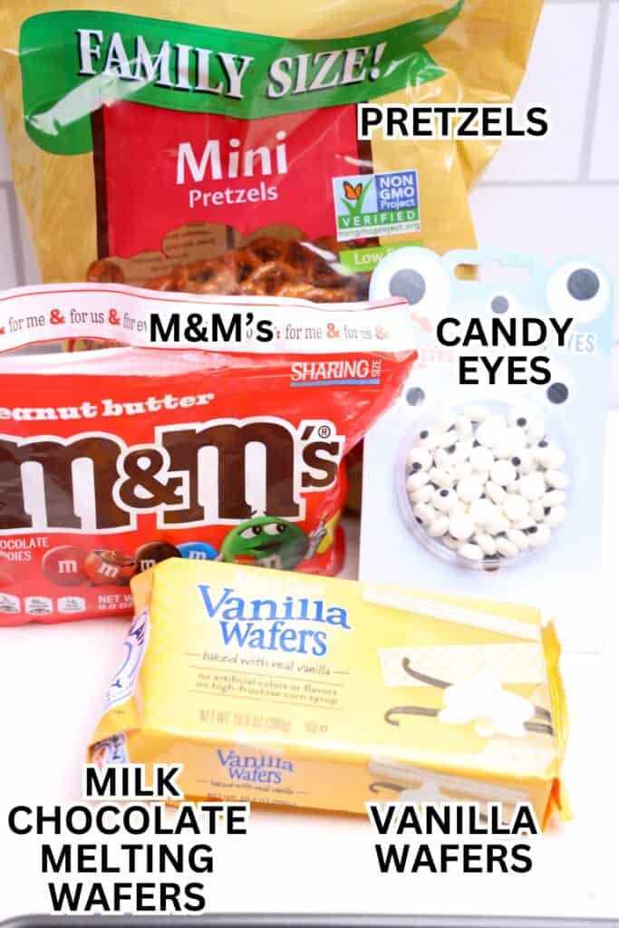 Ingredients for Christmas wafer reindeer cookies displayed on a counter, including a family-size bag of mini pretzels, peanut butter M&M’s, candy eyes, a package of vanilla wafers, and milk chocolate melting wafers. Each item is labeled to show what’s needed to assemble the festive treats.