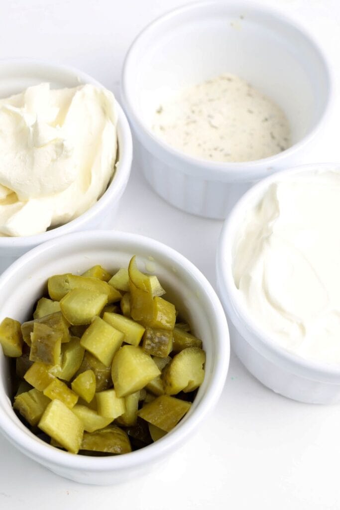 Four white ramekins filled with ingredients for dill pickle dip: chopped dill pickles, cream cheese, ranch seasoning mix, and sour cream. The ingredients are arranged on a clean white surface, ready to be mixed.