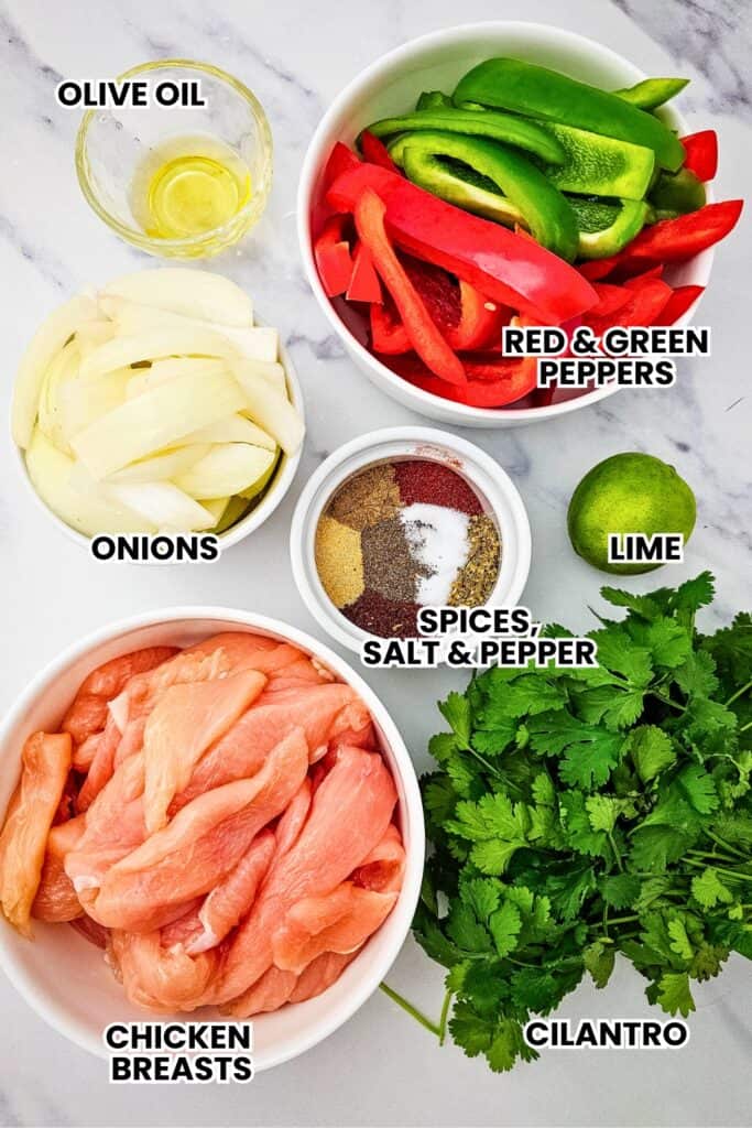 Ingredients for a fajitas sheet pan recipe arranged on a marble surface including sliced chicken breasts, red and green bell peppers, onions, olive oil, lime, fresh cilantro, and a small bowl of spices with salt and pepper. Each ingredient is labeled in bold text, showing everything needed to prepare baked chicken fajitas in the oven.