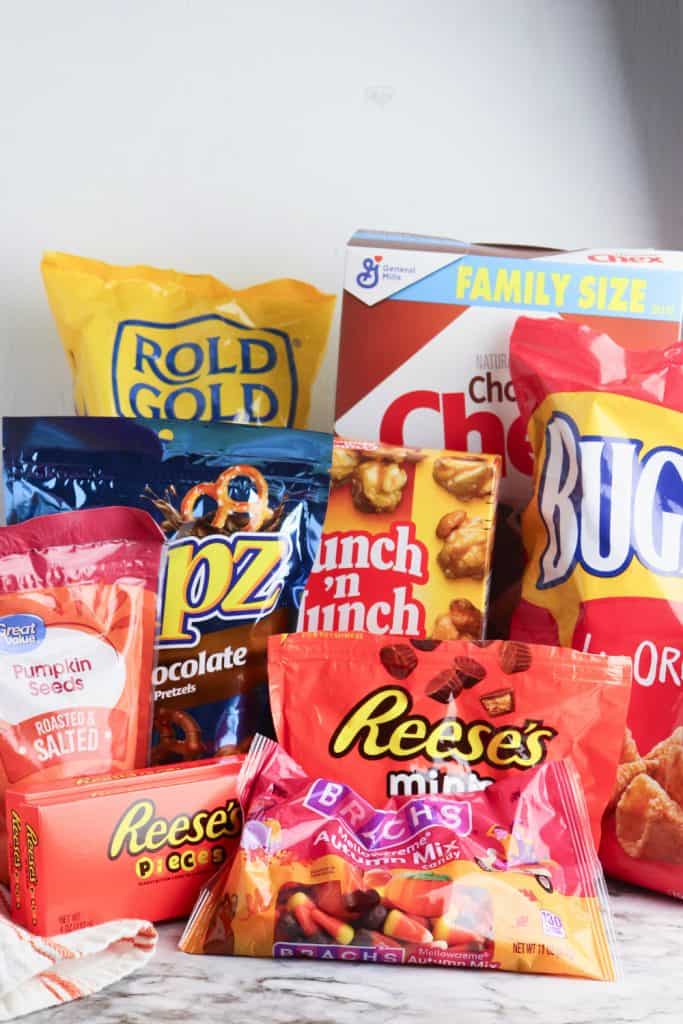 Assortment of packaged ingredients for a fall mix snack with candy corn, including Rold Gold pretzels, Chex cereal, Bugles, chocolate pretzels, Crunch 'n Munch, roasted pumpkin seeds, Reese’s Pieces, Reese’s Minis, and Brach’s Mellowcreme Autumn Mix candy, arranged on a marble surface against a white background.