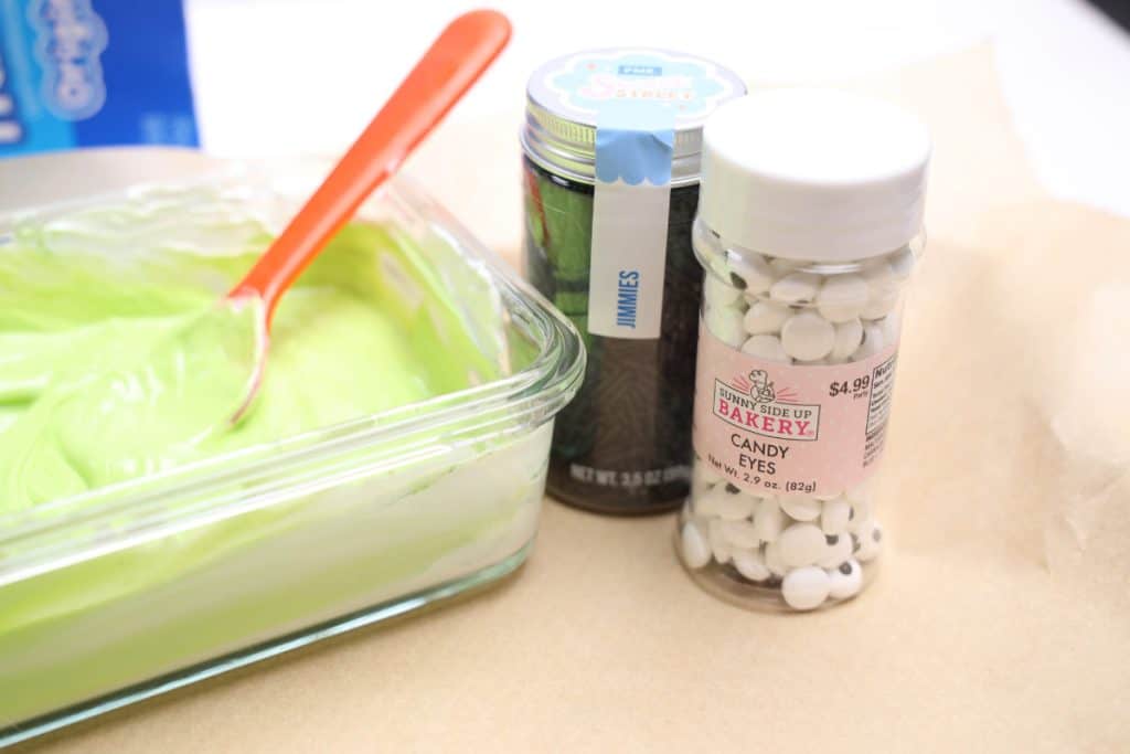 A close-up of Halloween treat ingredients including a bowl of green melted candy with a red spoon, a jar of black jimmies sprinkles, and a container of candy eyes from Sunny Side Up Bakery, all set on a sheet of parchment paper.