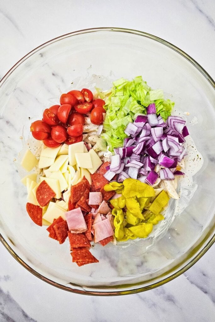 A large glass bowl filled with ingredients for hoagie pasta salad, including halved cherry tomatoes, shredded lettuce, diced red onion, sliced pepperoncinis, cubed provolone cheese, chopped ham, and pepperoni, all resting on a bed of penne pasta with creamy dressing. The ingredients are separated in colorful sections before mixing.