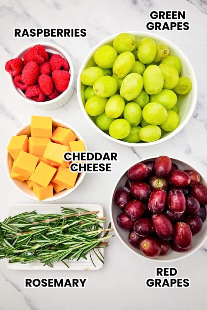 Ingredients for a holiday fruit and cheese platter arranged on a marble countertop, with white bowls of fresh raspberries, green and red grapes, cubed cheddar cheese, and rosemary sprigs.