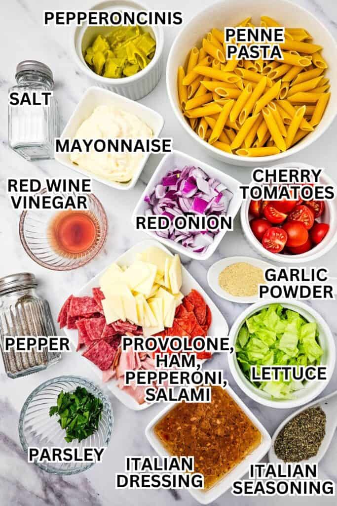 Overhead view of neatly arranged ingredients for an Italian sub pasta salad, including bowls of penne pasta, cherry tomatoes, lettuce, red onion, pepperoncinis, and sliced deli meats like ham, salami, and pepperoni. Also shown are provolone cheese, mayonnaise, red wine vinegar, Italian dressing, parsley, garlic powder, Italian seasoning, and salt and pepper, all labeled with bold text on a white marble surface.