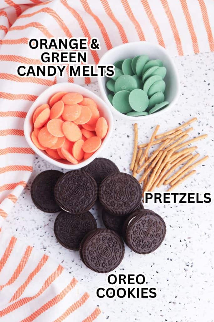 Flat lay of ingredients for a pumpkin-themed treat, including a bowl of orange and green candy melts, a pile of pretzel sticks, and a group of Oreo cookies. The items are arranged on a white speckled surface with an orange and white striped towel in the background.
