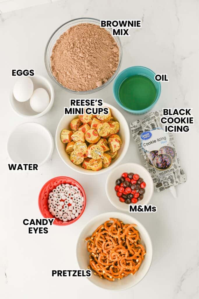 Flat lay of labeled ingredients for making brownie reindeer treats, including brownie mix, eggs, water, oil, Reese’s mini cups, candy eyes, red and brown M&M’s, pretzels, and black cookie icing. Each item is placed in a separate bowl or container on a white surface.