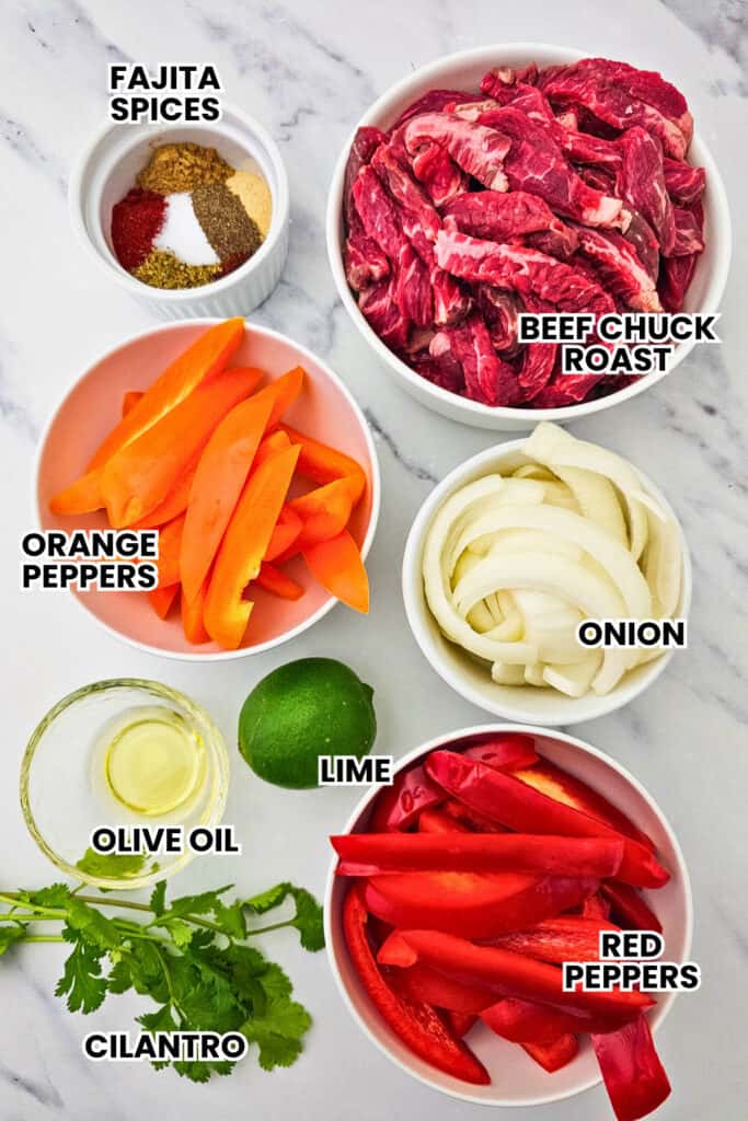 The ingredients for steak fajitas are arranged in small bowls on a marble surface. The ingredients are labeled as FAJITA SPICES, BEEF CHUCK ROAST, ORANGE PEPPERS, ONION, LIME, OLIVE OIL, RED PEPPERS and CILANTRO.