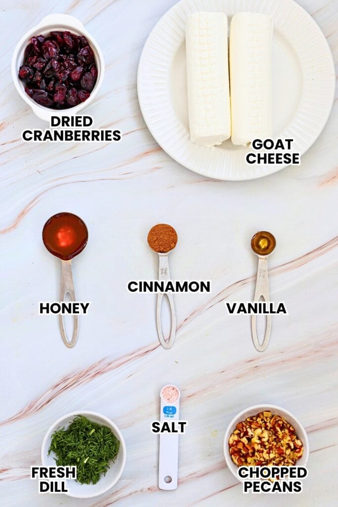 Ingredients for festive appetizers are arranged on a white marble surface. The labeled ingredients include dried cranberries, cream cheese, honey, cinnamon, vanilla, salt, fresh dill, and chopped pecans for mini pecan cranberry dill goat cheese balls.