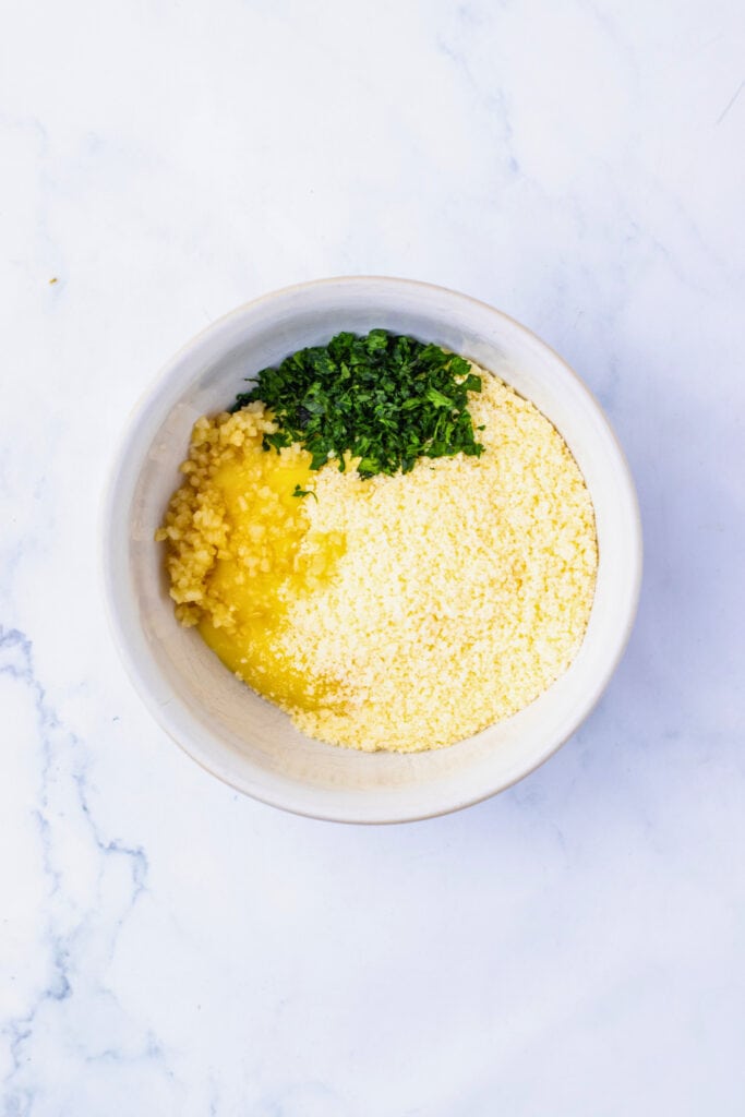 White mixing bowl filled with grated parmesan cheese, chopped parsley, minced garlic, and cream of chicken soup, ready to be stirred into a sauce for a baked chicken dish. The ingredients are neatly separated in the bowl before mixing.