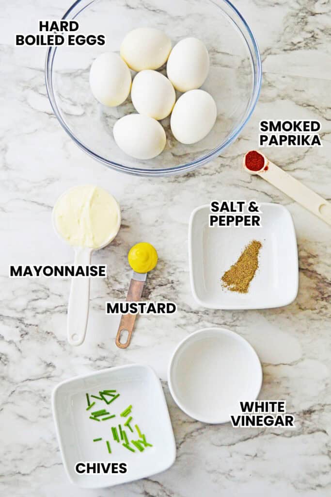 Ingredients for pumpkin deviled eggs in small bowls and measuring spoons arranged on a marble counter. Labels read "HARD BOILED EGGS" "SMOKED PAPRIKA" "SALT & PEPPER" "MAYONNAISE" "MUSTARD" "WHITE VINEGAR" and "CHIVES".