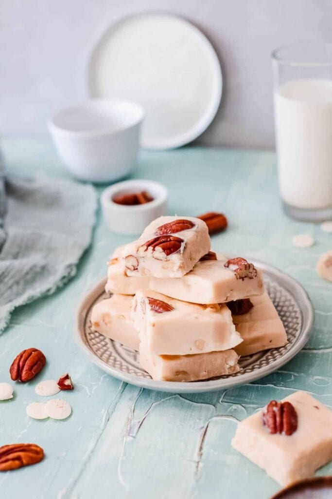 Creamy squares of Instant Pot butter pecan fudge stacked on a decorative plate, each piece studded with toasted pecans. The soft beige fudge sits on a pastel blue wooden surface, creating a festive and inviting Christmas dessert scene.