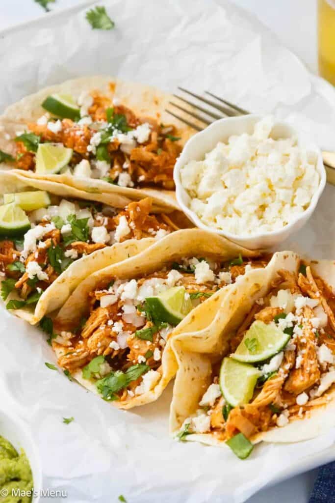 Instant Pot chicken tacos filled with shredded seasoned chicken and topped with diced onion, fresh cilantro, crumbled cotija cheese, and lime wedges. The tacos are arranged on a platter with extra cheese and garnishes served alongside.
