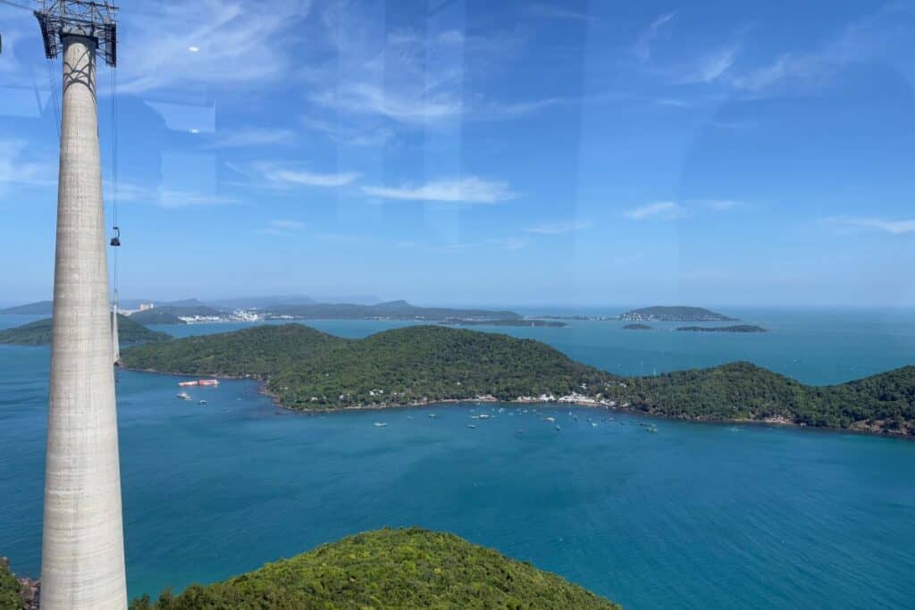 A panoramic view from the Hon Thom Cable Car in Phu Quoc, Vietnam, showing lush green islands surrounded by turquoise sea under a bright blue sky. A tall cable car support tower rises in the foreground, with boats dotting the coastline below.