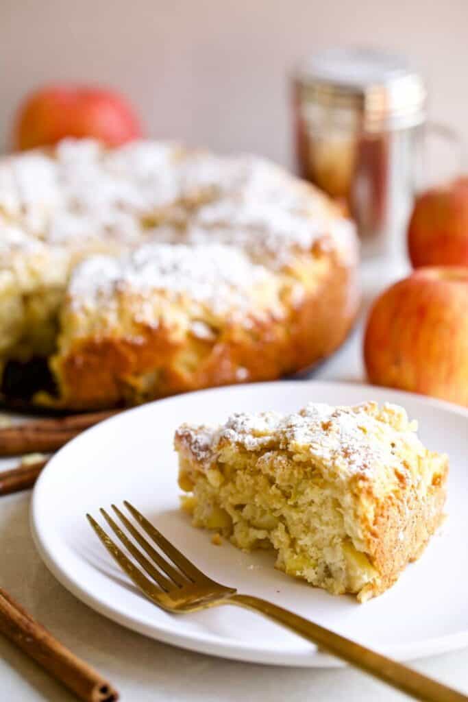 A slice of Italian apple cake, or Torta di Mele, sits on a white plate, showcasing its soft, moist crumb filled with tender apple chunks and lightly dusted with powdered sugar. The whole cake is visible in the background, making this rustic dessert a cozy and elegant choice for fall or Thanksgiving.