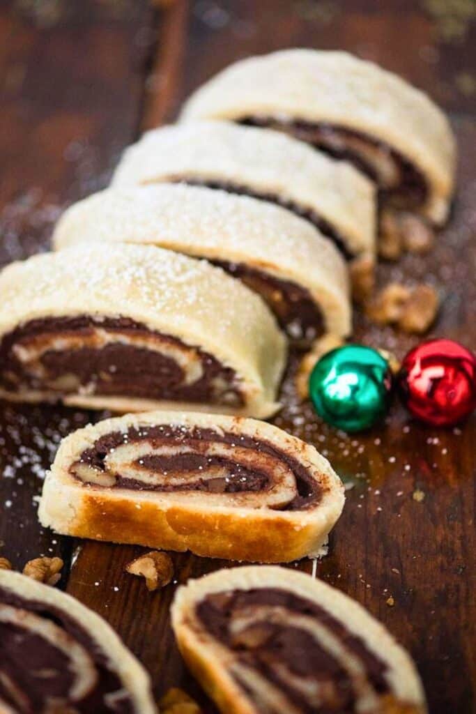 Sliced Italian chocolate roll cookies with a spiral of rich chocolate filling wrapped in golden, flaky pastry and dusted lightly with powdered sugar. The cookies are arranged on a rustic wooden surface with scattered walnuts and festive red and green jingle bells.