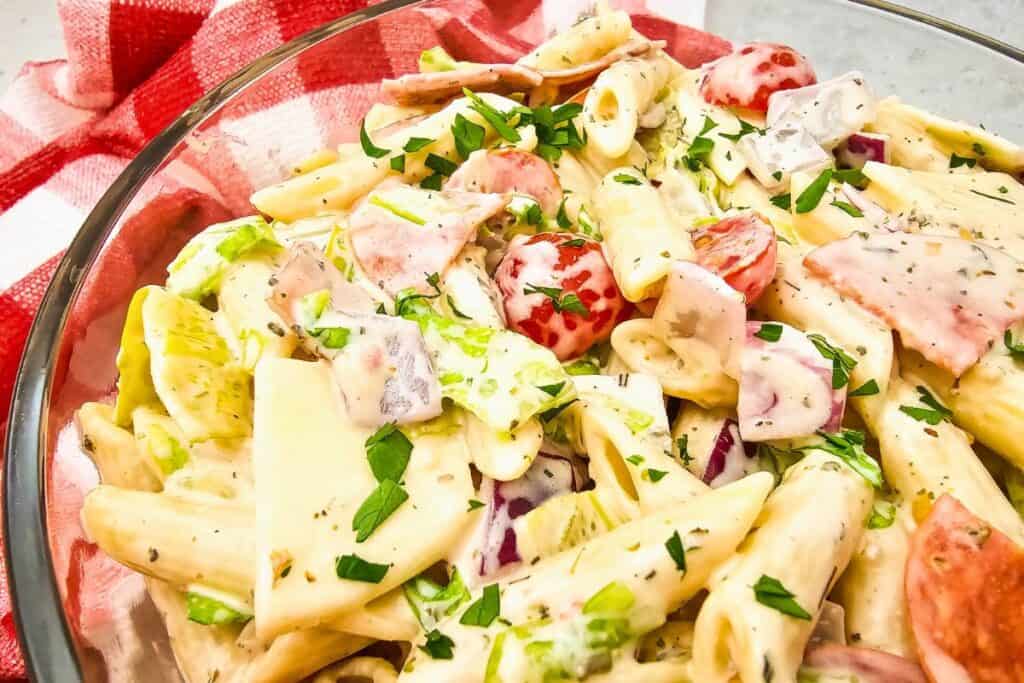 Close-up of a vibrant Italian hoagie pasta salad in a glass bowl, featuring creamy penne pasta mixed with lettuce, cherry tomatoes, red onions, sliced deli meats, provolone cheese squares, and a sprinkle of fresh parsley. The colorful dish is set against a red and white checkered cloth for a BBQ-style presentation.