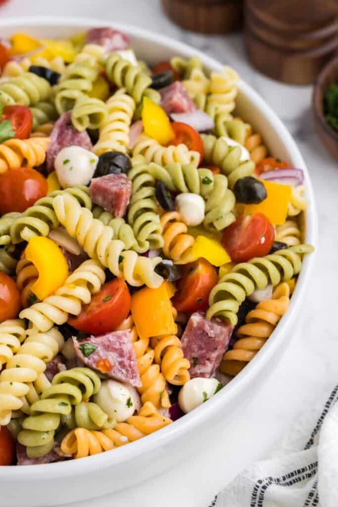 Large white bowl filled with Italian pasta salad made with tri color rotini cherry tomatoes black olives yellow bell peppers red onion mozzarella balls and cubes of salami. The colorful pasta salad is tossed with herbs and dressing.