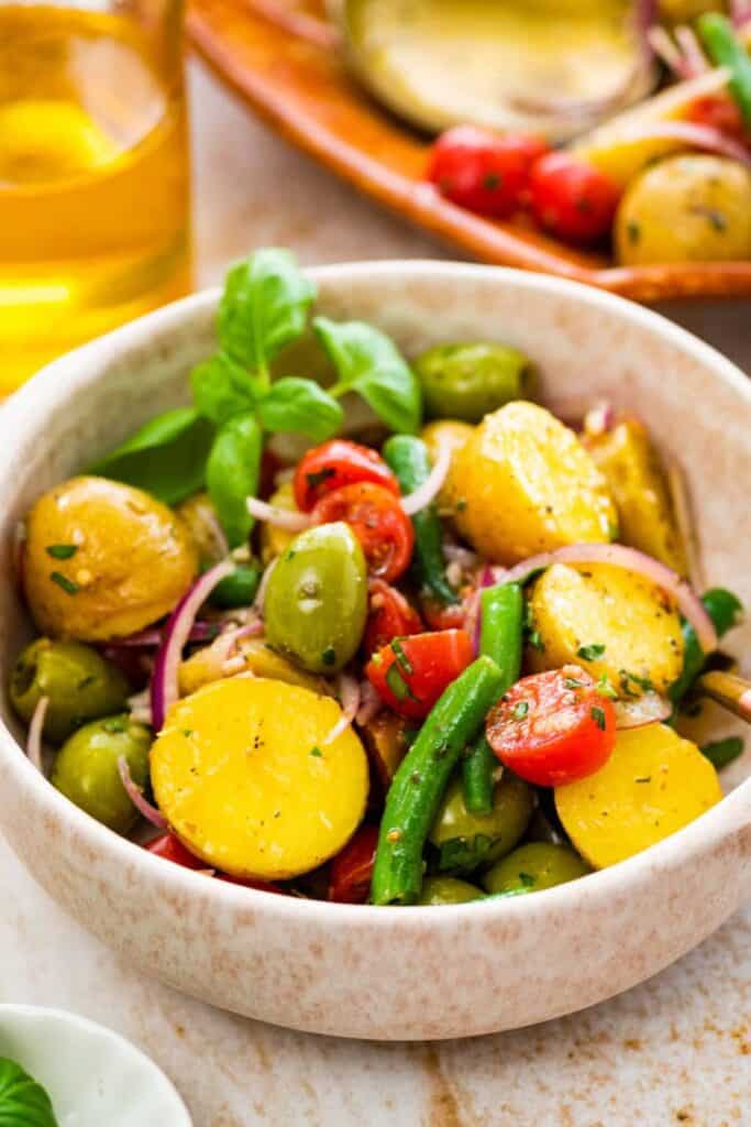 A vibrant Italian potato salad served in a rustic beige bowl, featuring halved yellow potatoes, green beans, cherry tomatoes, green olives, and thinly sliced red onion, tossed in a light herb vinaigrette and garnished with fresh basil leaves.
