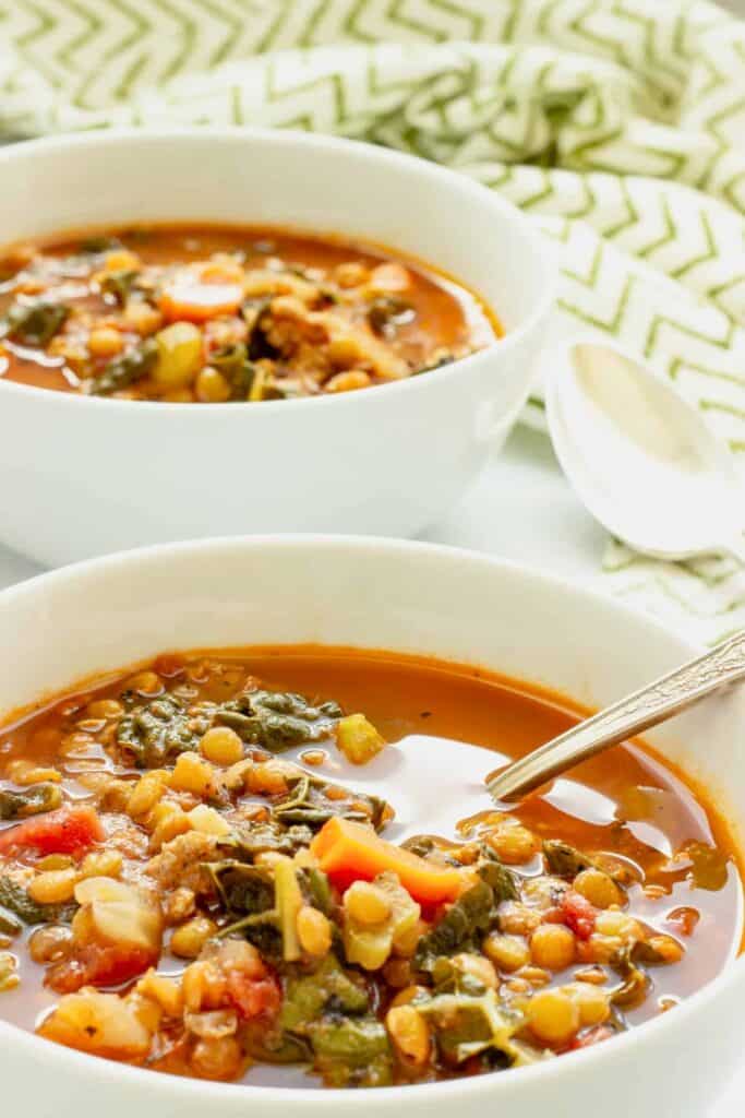 Italian sausage and lentil soup with leafy greens tomatoes carrots and vegetables is served in a rich tomato broth. Two white bowls showcase the hearty soup ready for easy cold weather dinner.