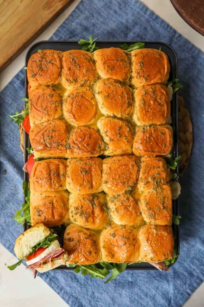 A tray of golden-brown Italian sliders made with soft, herb-buttered rolls, stacked with layers of deli meats, cheese, tomatoes, and fresh greens ready to serve, with one sandwich pulled out to show the colorful filling.