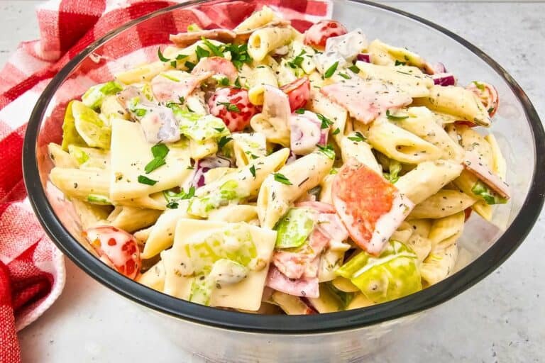 A clear glass bowl filled with creamy Italian sub pasta salad, featuring penne pasta, sliced deli meats, provolone cheese squares, cherry tomatoes, lettuce, red onion, and chopped parsley. The salad is richly coated in a seasoned dressing and placed on a white surface next to a red and white checkered towel, giving it a fresh, picnic or potluck style appeal.