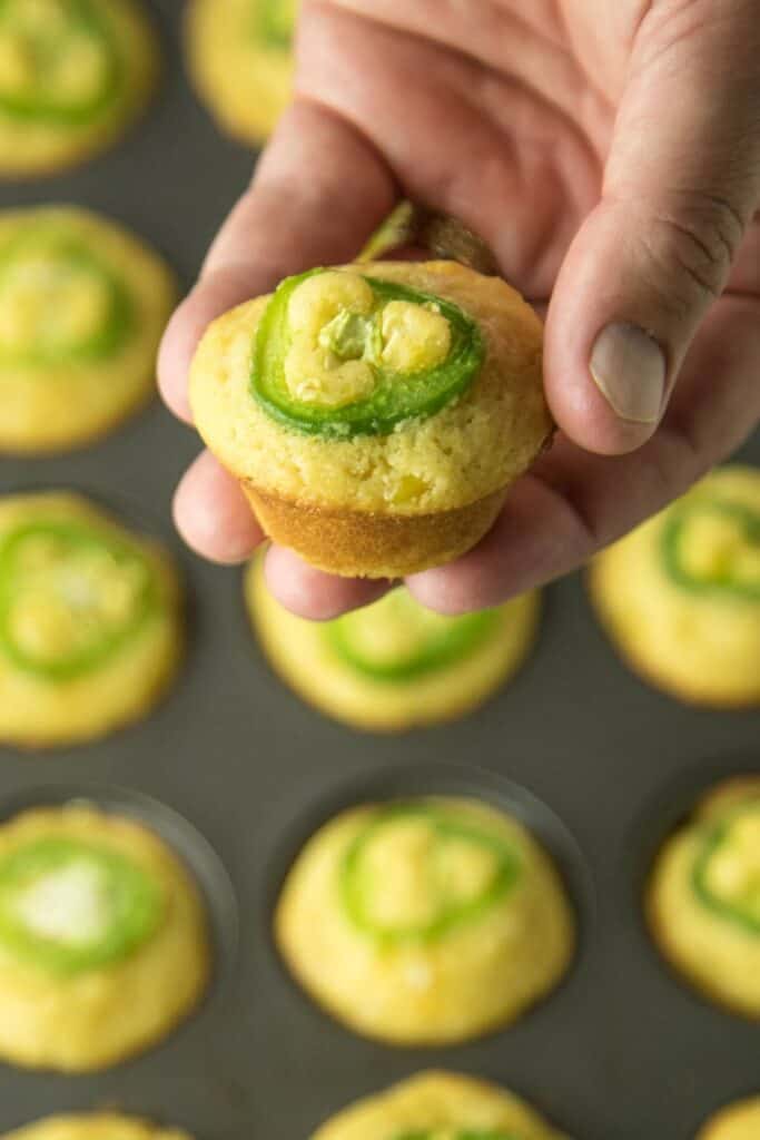 Close-up of a hand holding a mini cheesy jalapeño cornbread muffin, topped with a fresh jalapeño slice. More golden muffins sit in a dark muffin tin below, each with melted cheese and spicy jalapeño baked into the top.