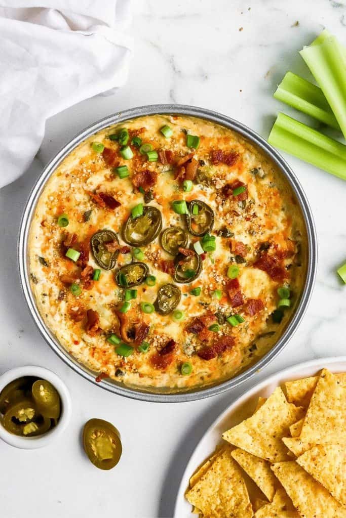 Creamy jalapeño popper dip in a metal dish, topped with sliced jalapeños, crispy bacon, and chopped green onions, served with celery sticks and tortilla chips. A spicy and cheesy game day finger food sized for sharing.