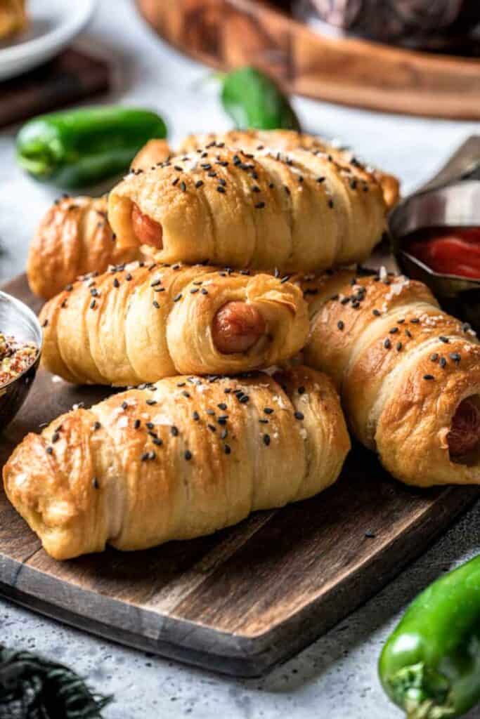 Golden-brown bagel dog bites wrapped in flaky dough, sprinkled with black sesame seeds and sea salt, served on a wooden board with dipping sauces and fresh jalapeños nearby.