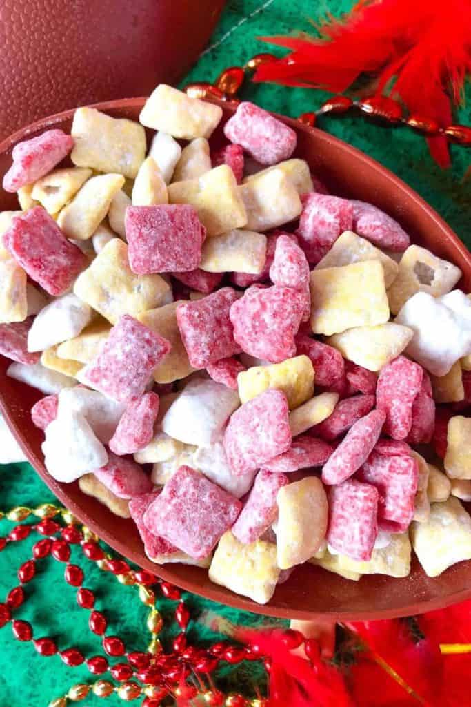 A festive bowl filled with red, yellow, and white powdered muddy buddies, arranged to match the Kansas City Chiefs' team colors. The colorful snack sits on a green background decorated with shiny red and gold beads and red feathers, evoking a game day or holiday celebration.
