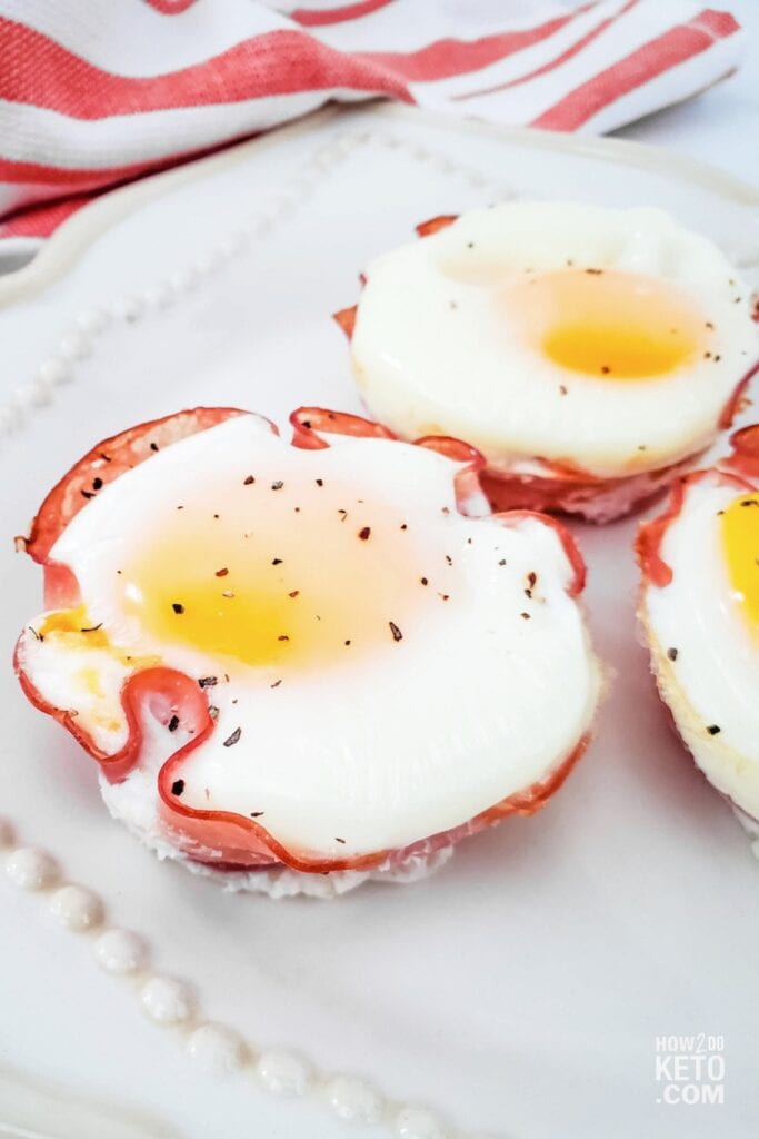 Three keto egg cups made with baked eggs set inside crispy ham slices and topped with black pepper on a white plate.