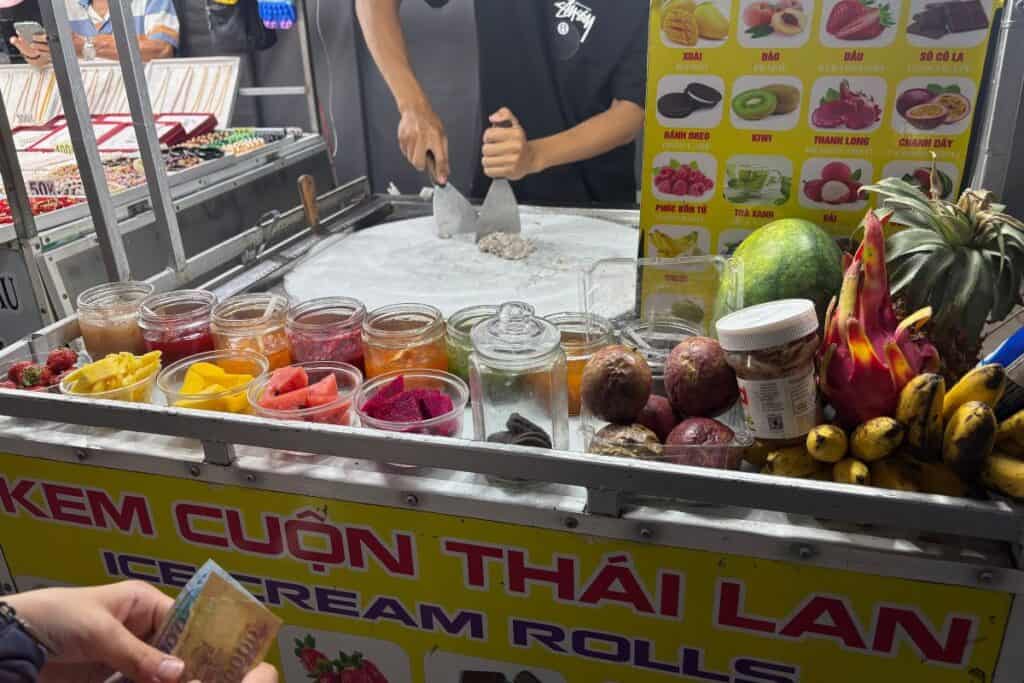 A colorful rolled ice cream stand at Phu Quoc Night Market, featuring jars of fresh fruit like mango, dragon fruit, and watermelon, with a vendor preparing rolled ice cream on a cold round surface.
