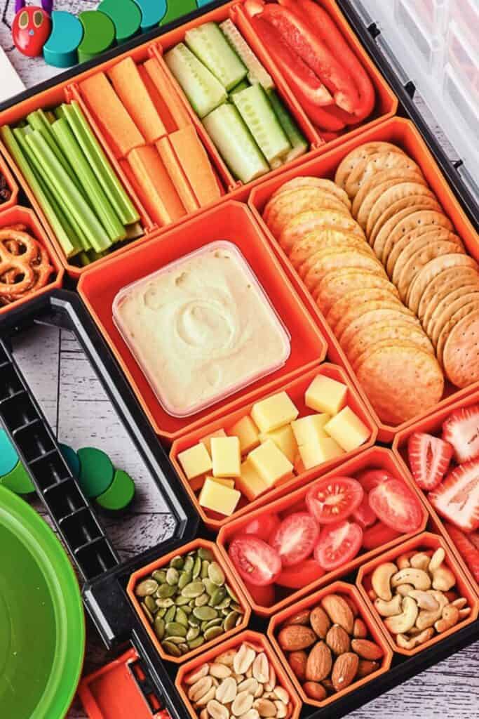 Colorful kids charcuterie board arranged in a compartmentalized snack box with sliced cucumbers, carrot sticks, celery, and red bell pepper strips. The box also includes round crackers, cheese cubes, hummus dip, cherry tomatoes, strawberries, almonds, cashews, pumpkin seeds, and peanuts, creating a balanced mix of fresh produce and crunchy snacks.