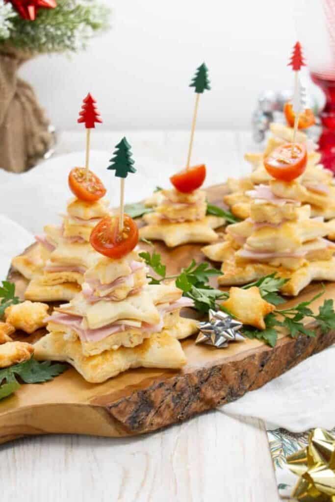 Wooden cutting board with Christmas tree appetizers made out of puff pastry, cheese and topped with a cherry tomato. 