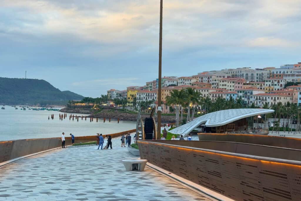 View from the Kiss Bridge at sunset, Phu Quoc Island, showing people strolling along the modern patterned walkway with warm lighting. In the background, colorful hillside buildings of Sunset Town overlook the ocean, with anchored boats and a soft, cloudy sky completing the peaceful coastal scene